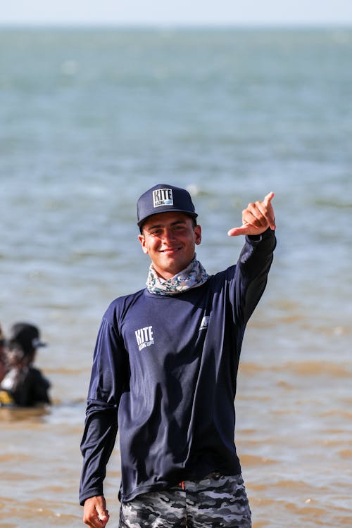 Man in a Cap Standing by a Sea and Making Hand Gesture