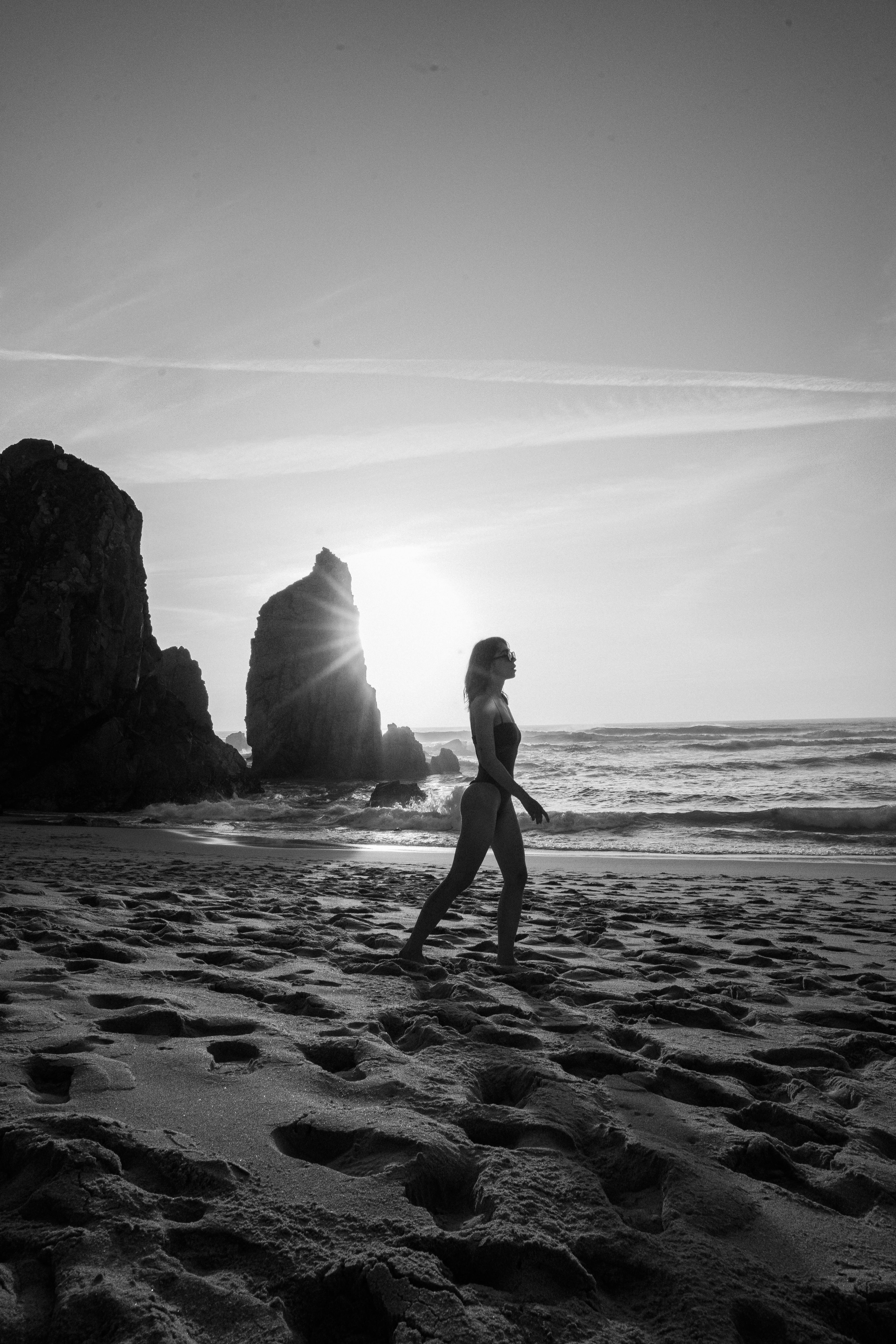 Black and white silhouette of woman walking along a rocky beach at sunset, creating a dramatic scene.