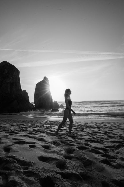 Black and white silhouette of woman walking along a rocky beach at sunset, creating a dramatic scene.