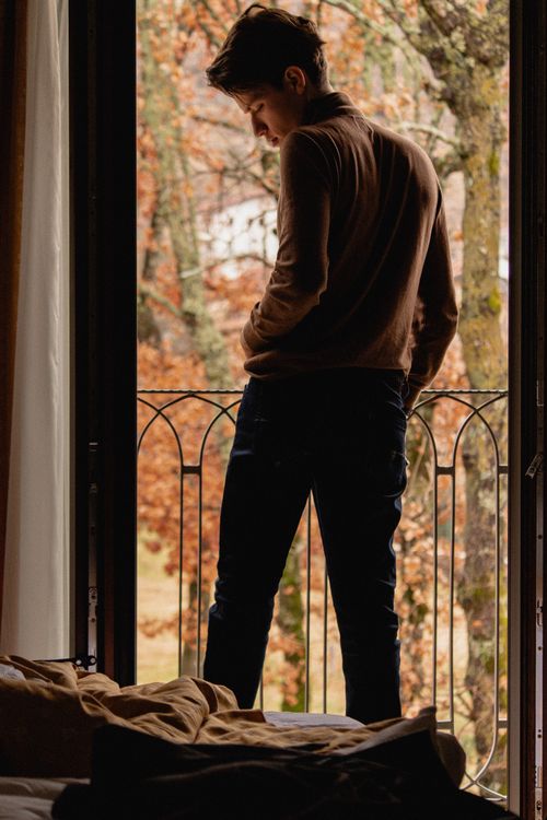 Free Man in brown attire gazes thoughtfully outside through an open doorway, surrounded by autumn trees. Stock Photo