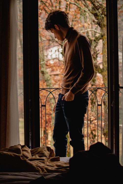 Free A man in brown sweater gazing outside from a balcony during fall, framed by a glass door. Stock Photo