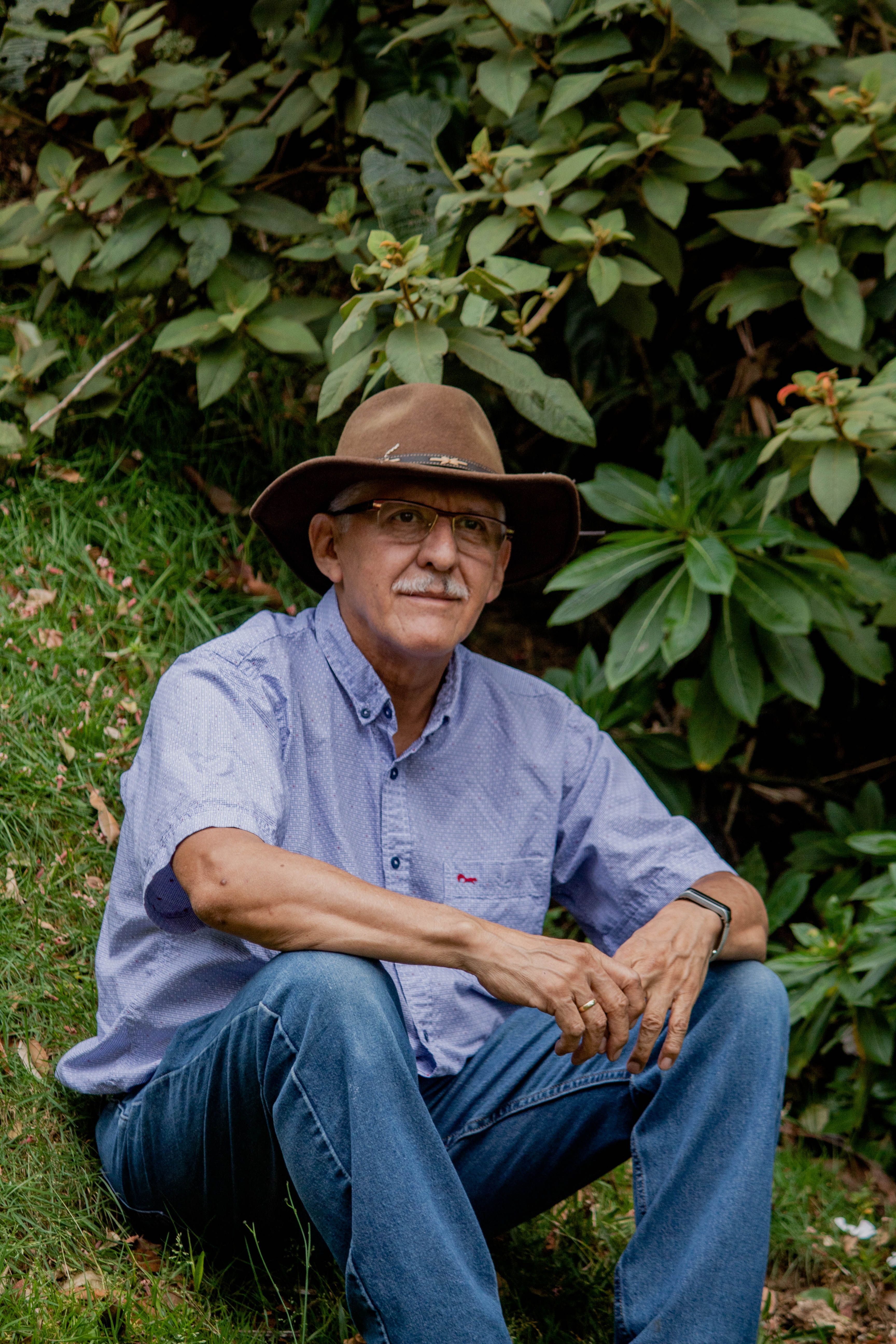 A senior man wearing a cowboy hat and eyeglasses sitting on grass in a rural outdoor setting.