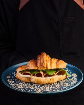 Close-up of a croissant sandwich served on a blue plate, presented by a person in a black apron.