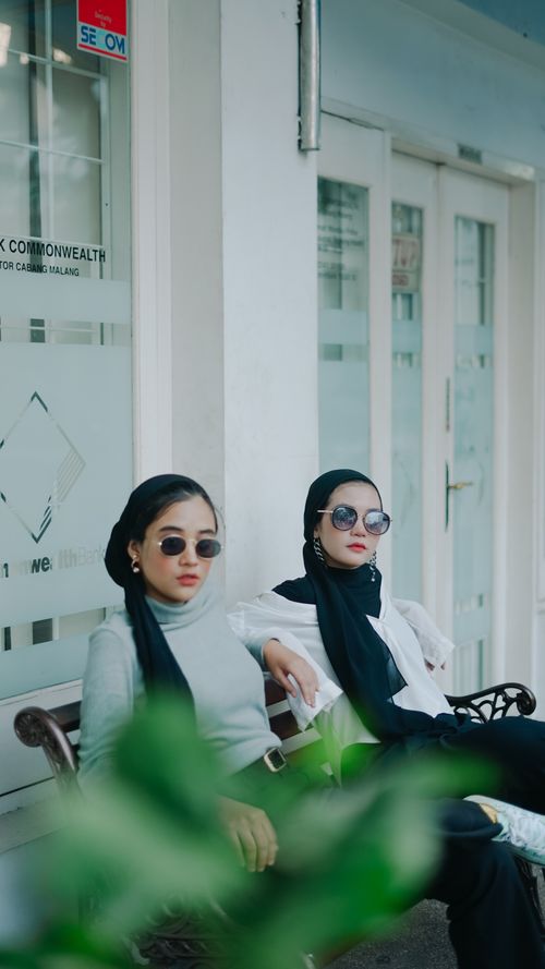 Free Two fashionable women wearing hijabs and sunglasses seated outdoors. Stock Photo