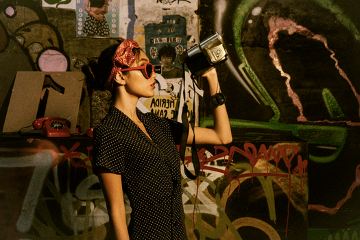 Woman in fashionable polka dot dress posing with video camera indoors by graffiti.