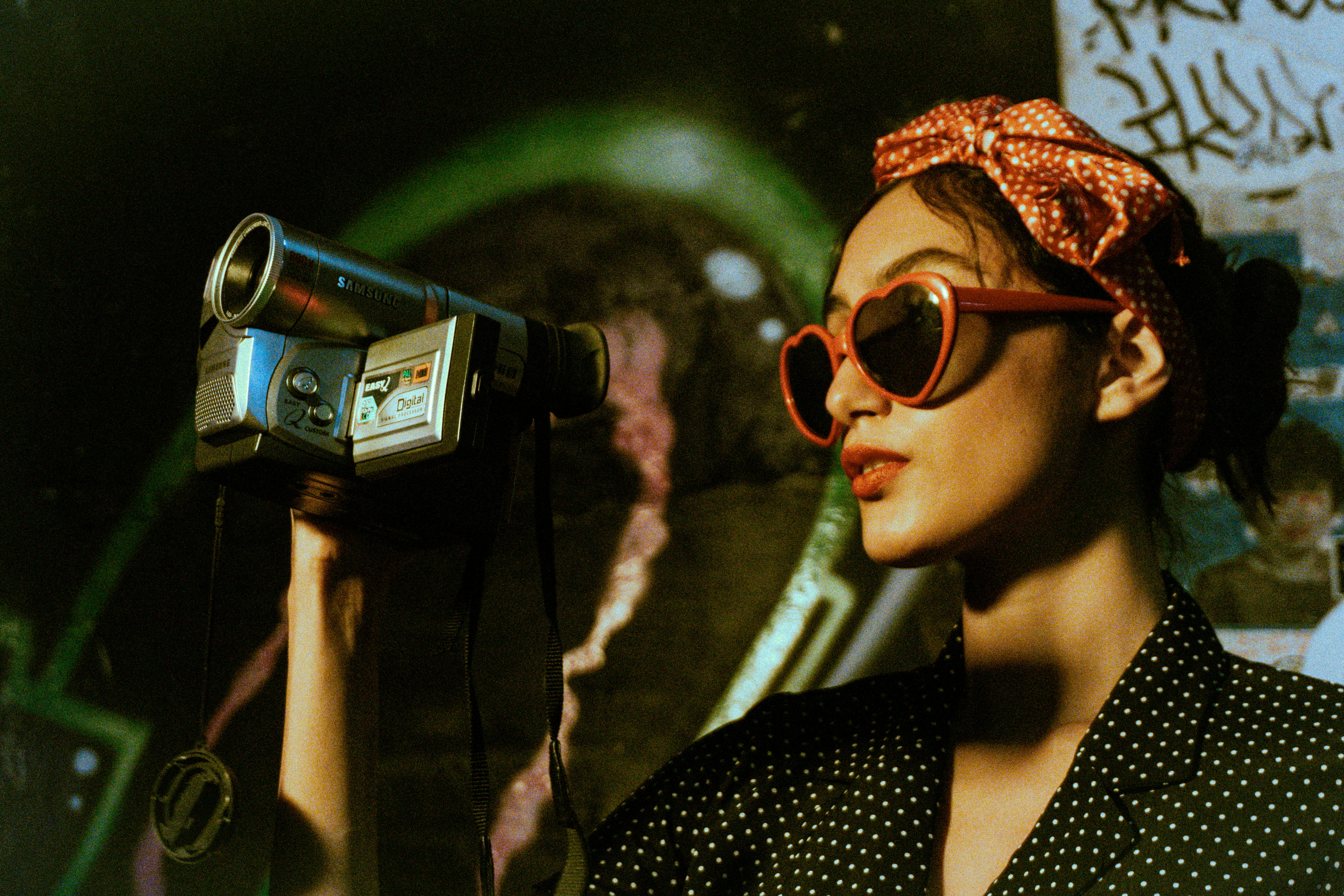 a woman in black polka dots shirt wearing eyeglasses while holding a video camera