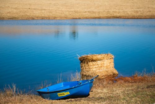 Free A serene rural landscape featuring a blue boat and hay bale by a calm river. Stock Photo