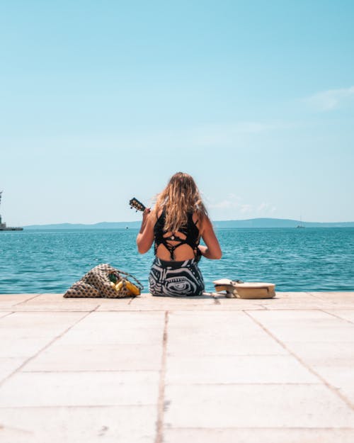 Free A Person Playing a Guitar while Sitting by the Seaside Stock Photo