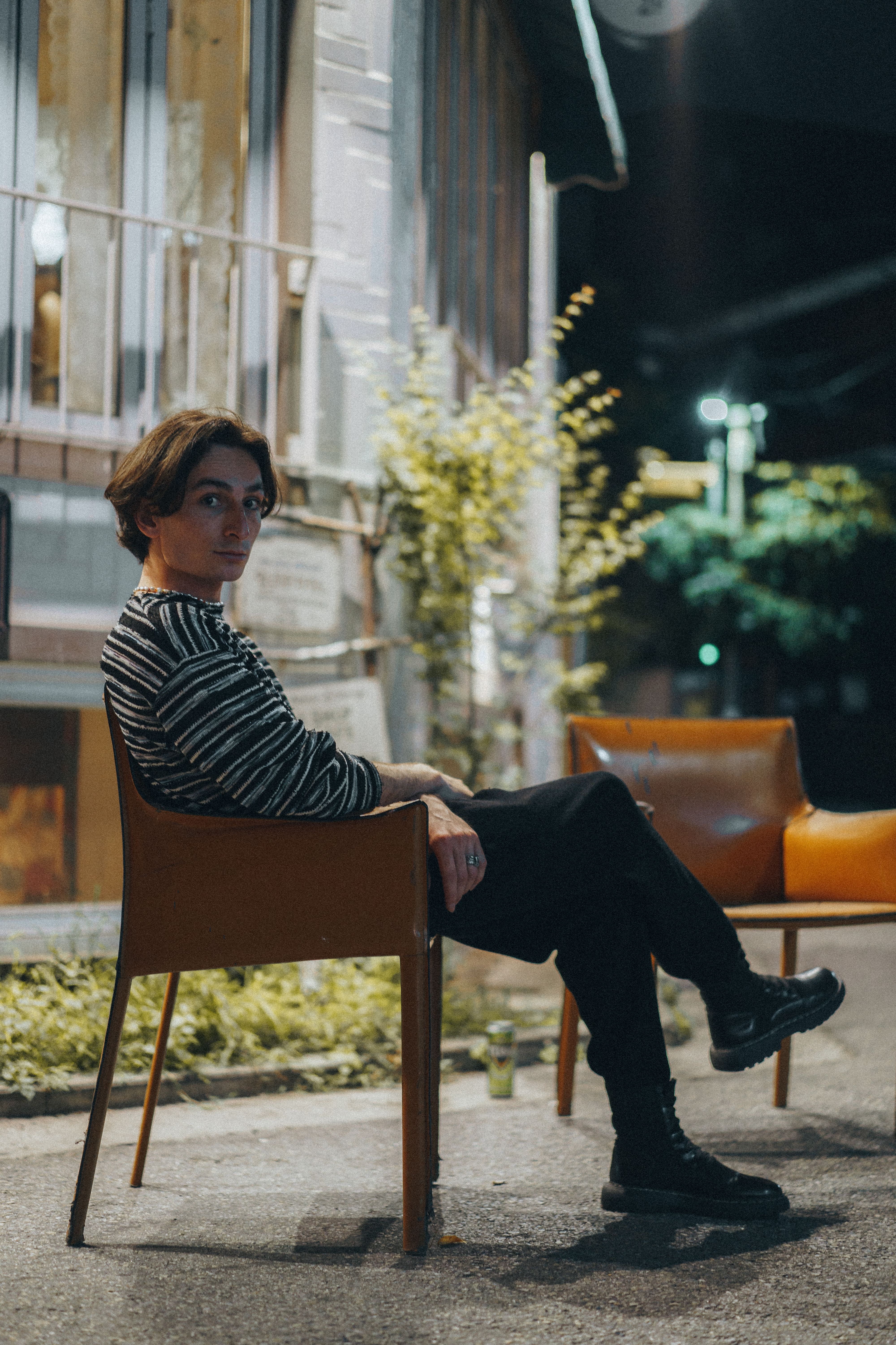 A stylish man in a striped shirt sitting outside at night, city backdrop.