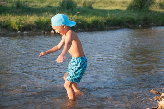 A young boy wearing a cap and shorts enjoys a playful moment in a shallow river on a bright summer day.