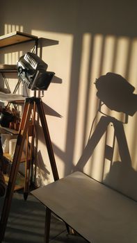 A vintage tripod light casting artistic shadows in a sunlit studio room with shelves.
