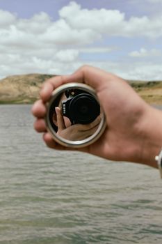 A hand holding a mirror reflects a camera with a scenic lake backdrop.