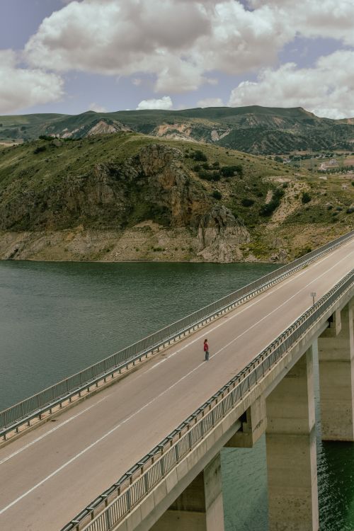 Free A lone person walks on a bridge over a river with scenic mountains in the background. Stock Photo
