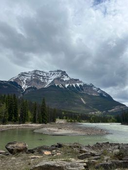 Breathtaking view of snow-capped mountains in Canada with river and trees under a cloudy sky.