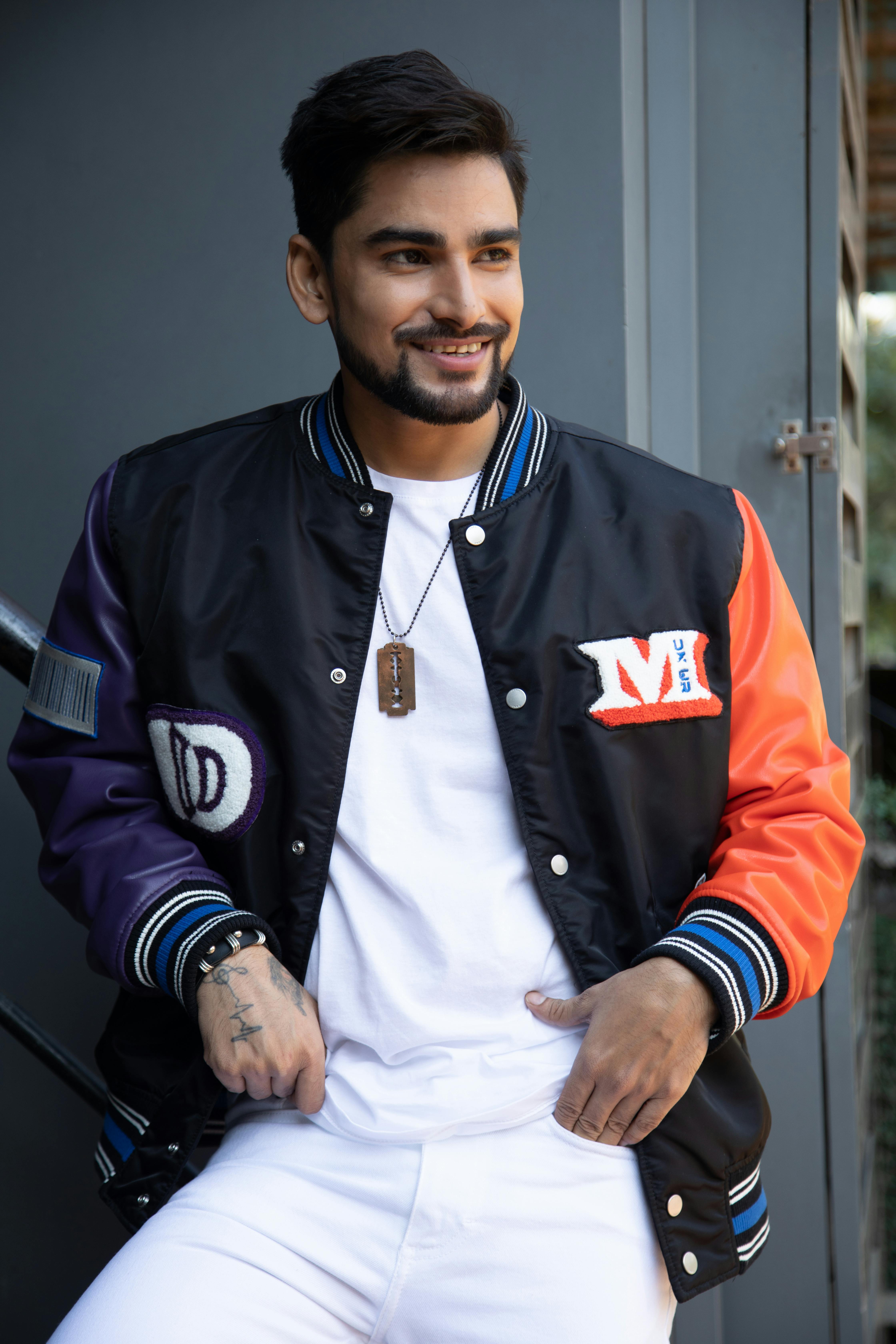 Portrait of a young stylish man wearing a colorful baseball jacket, confidently posing and smiling outdoors.