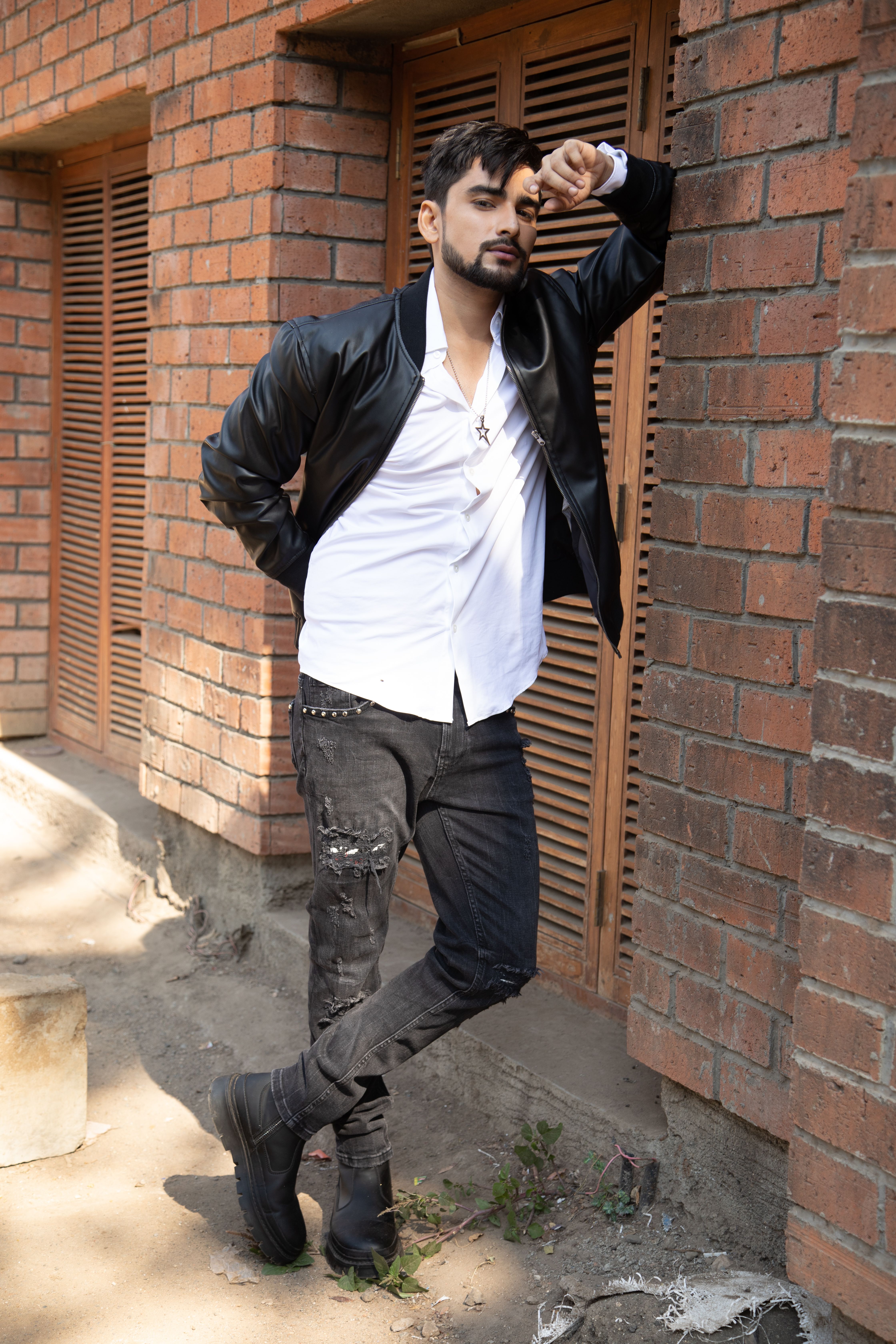 A stylish man in a leather jacket leans against a brick wall, showcasing a modern fashion pose.