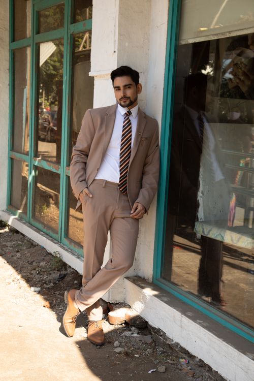 Free A stylish man in a tan suit leans against a window outdoors, showcasing modern fashion. Stock Photo