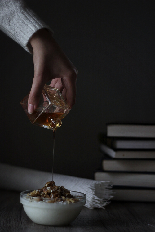 Free A hand pours honey over yogurt topped with nuts, set against a stack of books, evoking a cozy, dark ambiance. Stock Photo