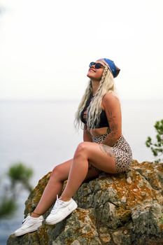 Blonde woman with tattoos smiling on a rock by the sea, enjoying the summer day.