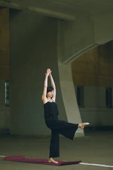 A woman in a yoga stance with arms raised and one leg lifted, in an indoor setting.