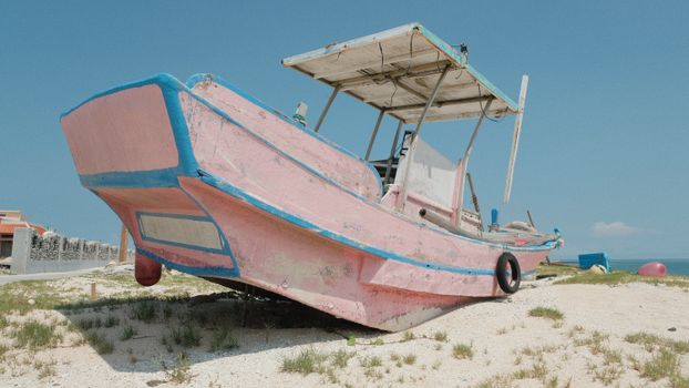 An abandoned pink wooden fishing boat rests on a sandy shore in Taiwan, under a clear blue sky.