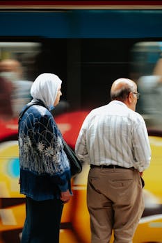 Elderly couple waiting at a busy station with a train passing by, capturing a moment in motion.