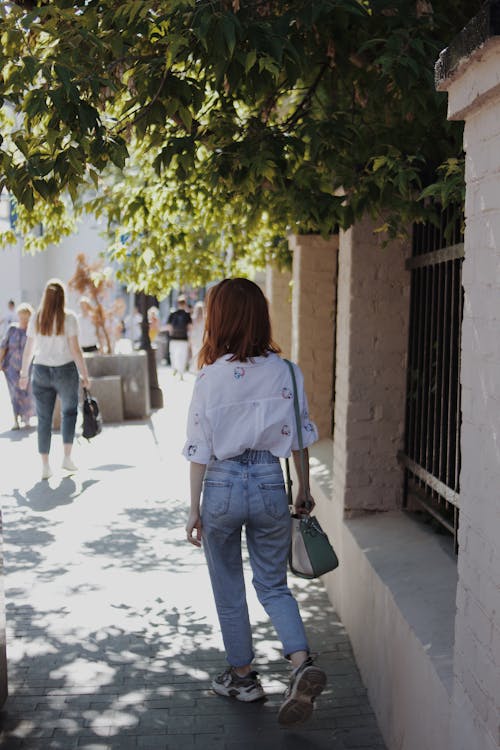 A Woman in a White Top and Denim Pants Walking at the Sidewalk