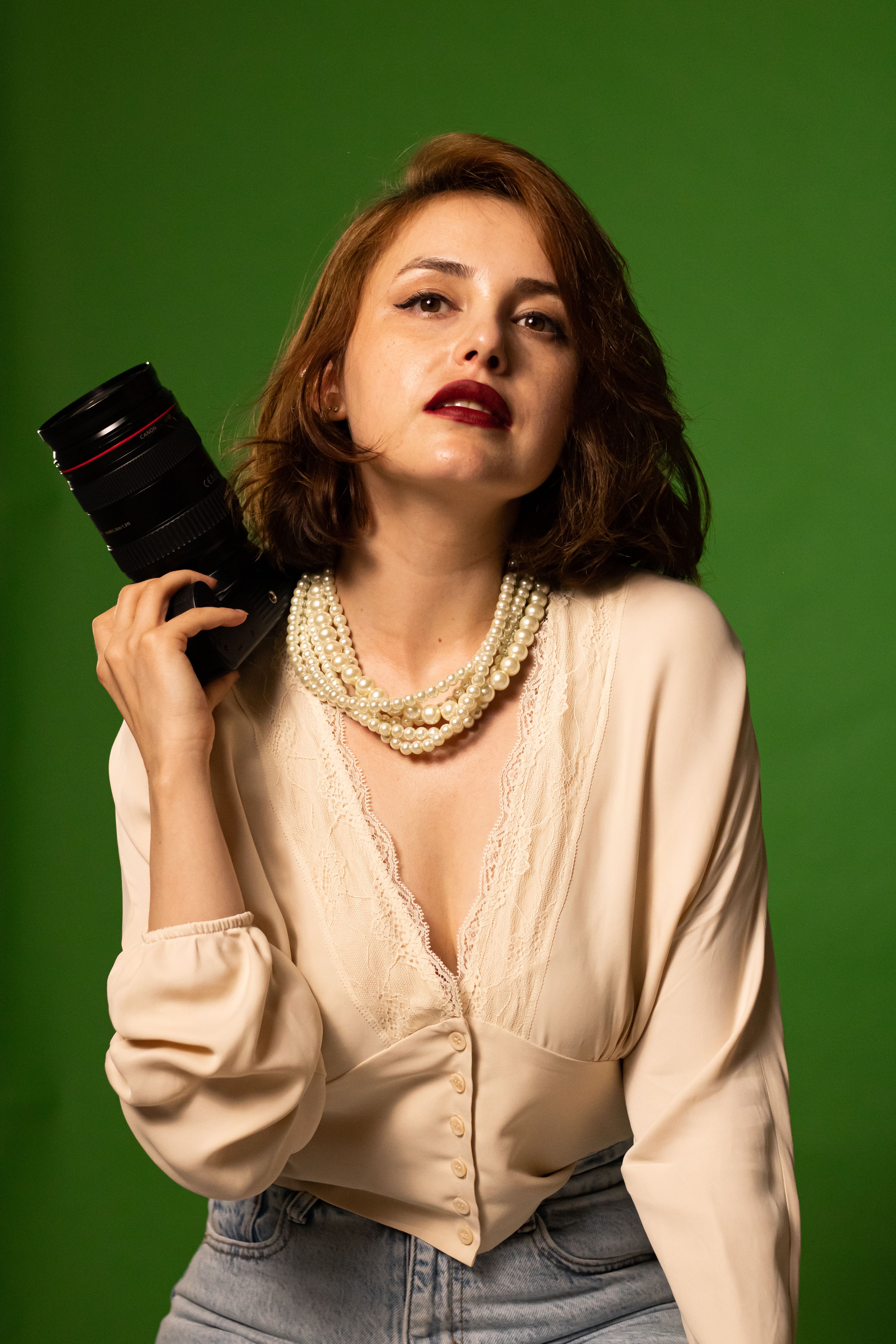 A stylish woman in beige blouse holding a camera, standing in front of a green studio backdrop.