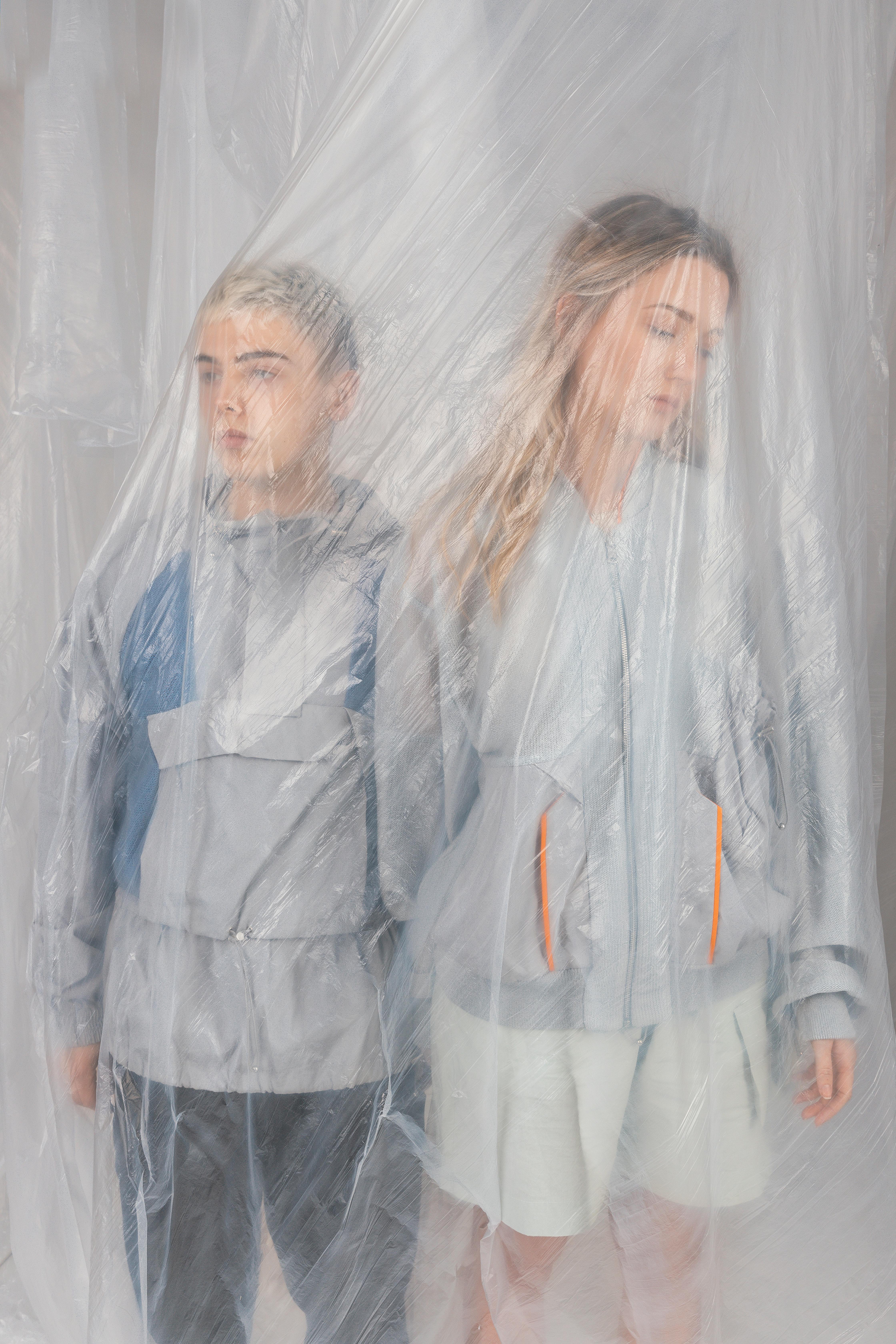 Contemporary fashion portrait of two adults behind translucent plastic, showcasing minimalistic style.