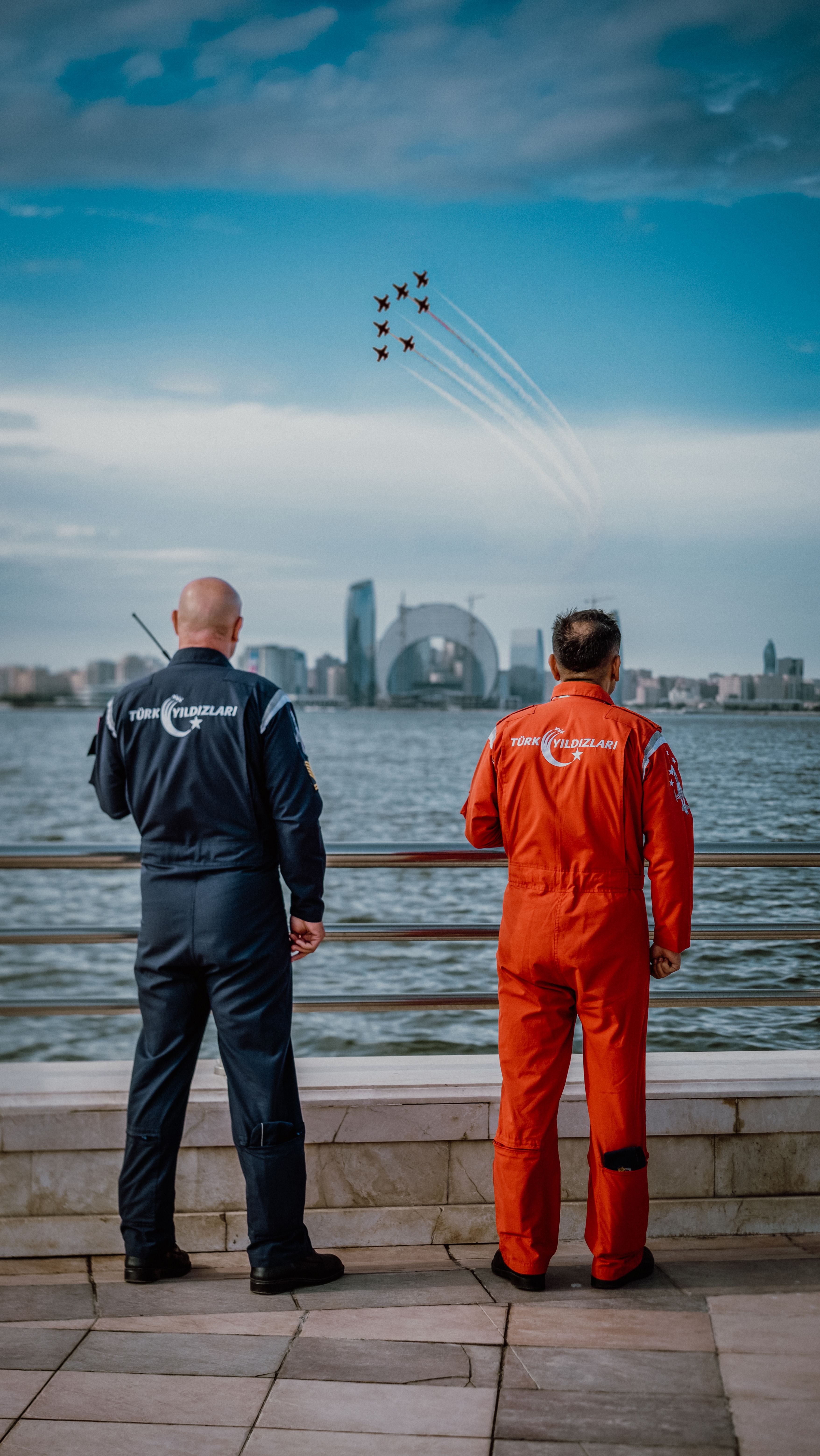 Pilots in vibrant suits watch airplanes performing aerial stunts over Baku's cityscape.