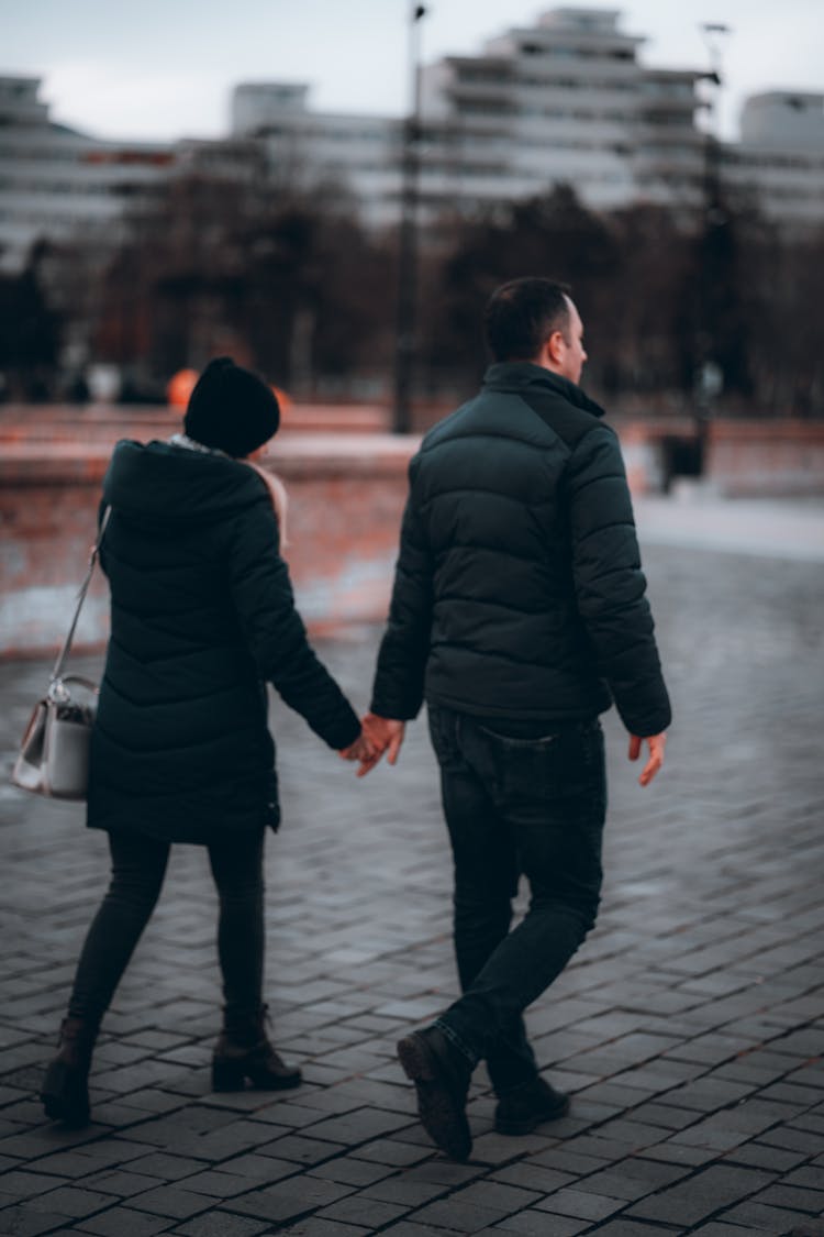 A Couple In Black Puffer Jackets Walking On The Street While Holding Hands