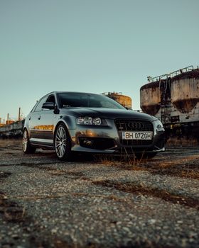 A luxury Audi sedan parked on pavement with industrial tanks in the background, captured during sunset.
