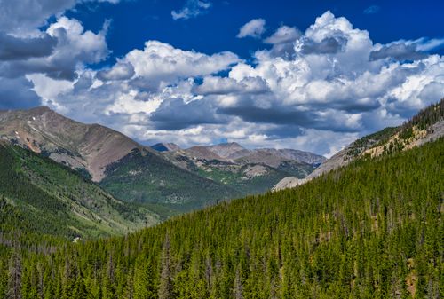 Free Explore the breathtaking view of mountains and forests under a vibrant blue sky with fluffy clouds. Stock Photo