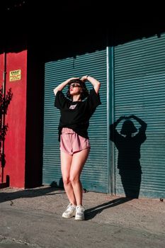 Stylish woman in pink shorts and black top posing on a sunny street with dramatic shadows.