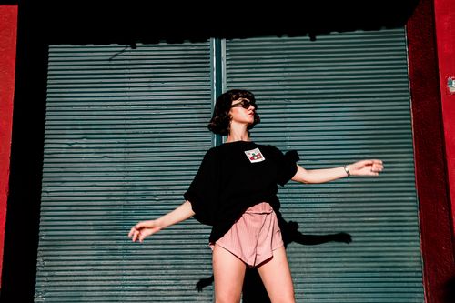 Free Stylish woman in sunglasses and vibrant shorts posing by a roll-up door on a sunny day. Stock Photo
