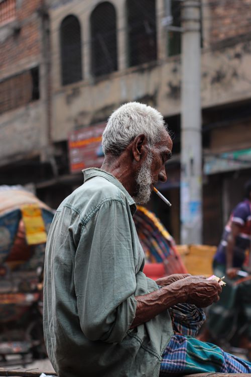 Free A senior man with a cigarette sits outdoors on a bustling city street, reflecting a busy lifestyle. Stock Photo
