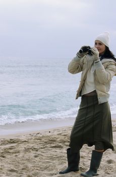 Woman in winter attire using binoculars on a beach by the ocean, looking at the waves.
