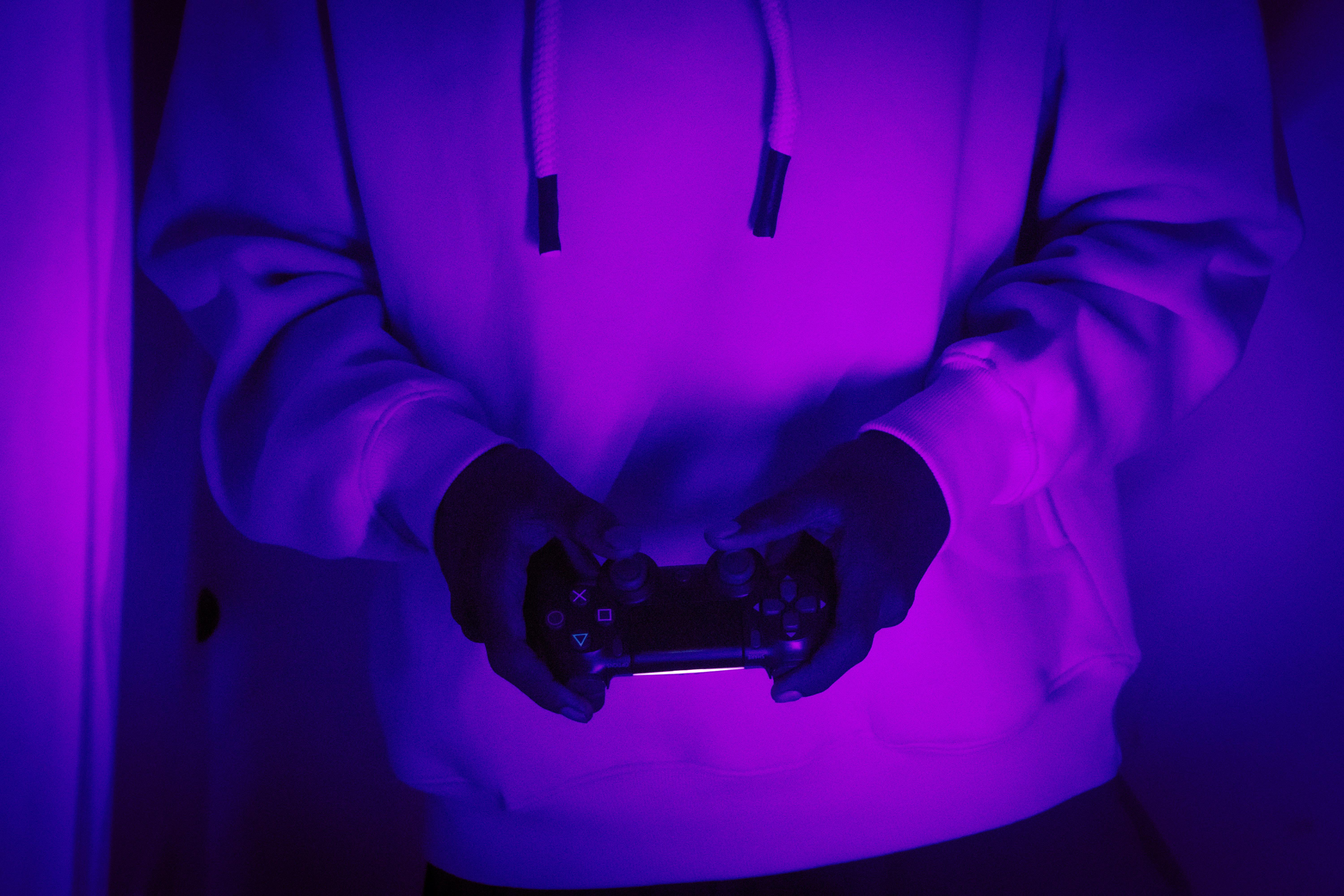 A person in a hoodie holds a game controller under neon purple light.