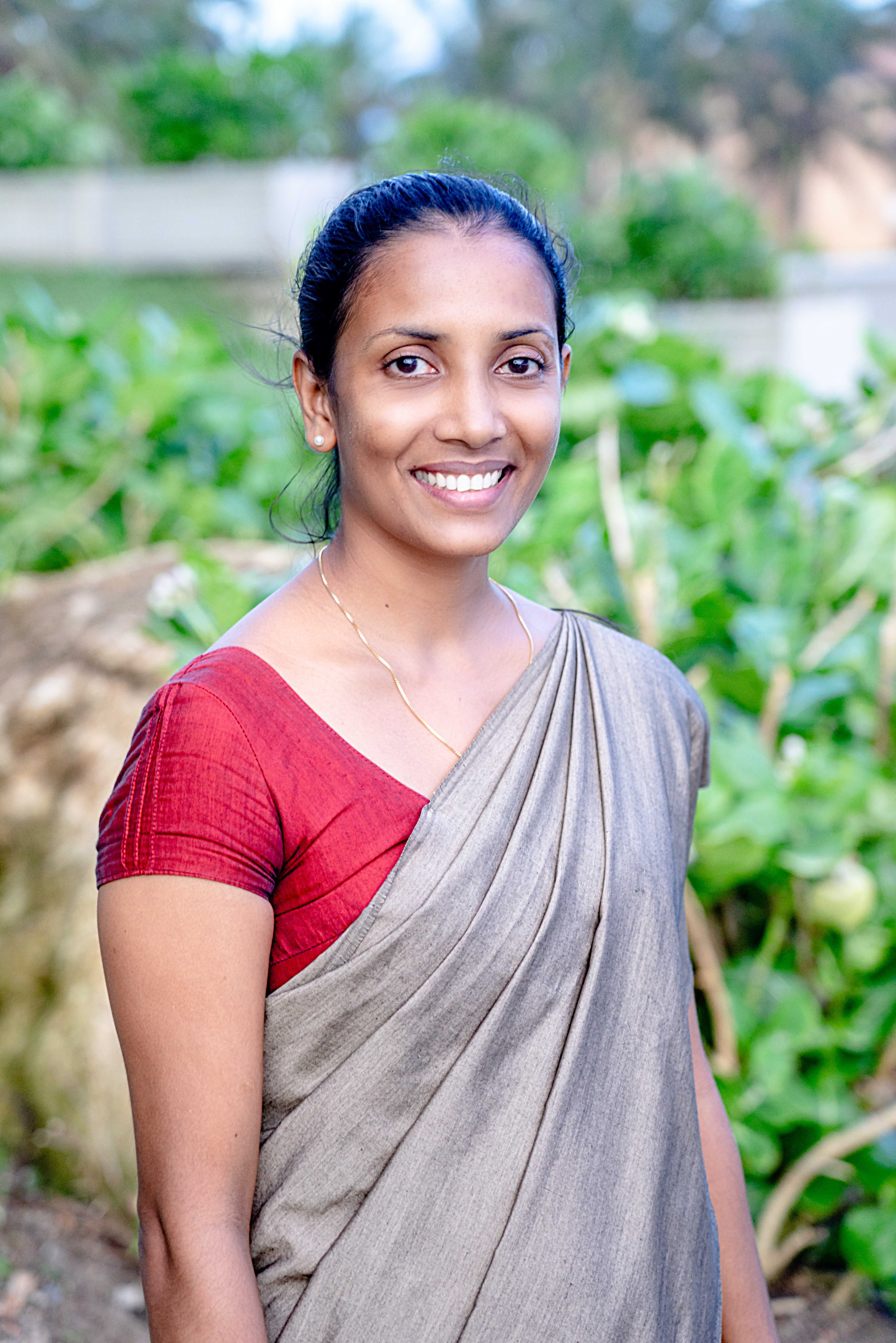 Portrait of a smiling woman in a traditional sari standing outdoors. The lush green background adds vibrancy.