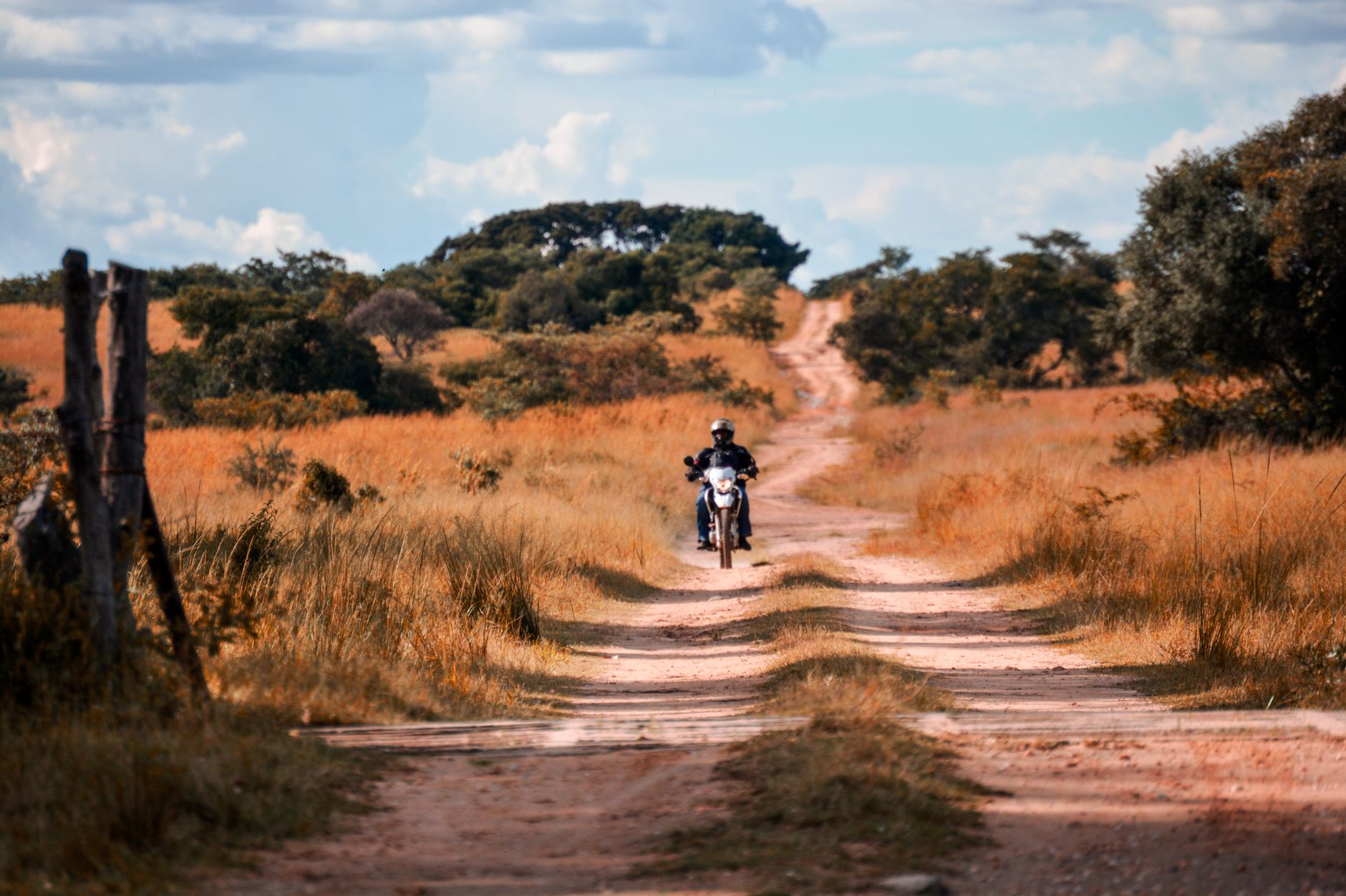 A motorcyclist rides through a picturesque dirt road in a rural savannah landscape.