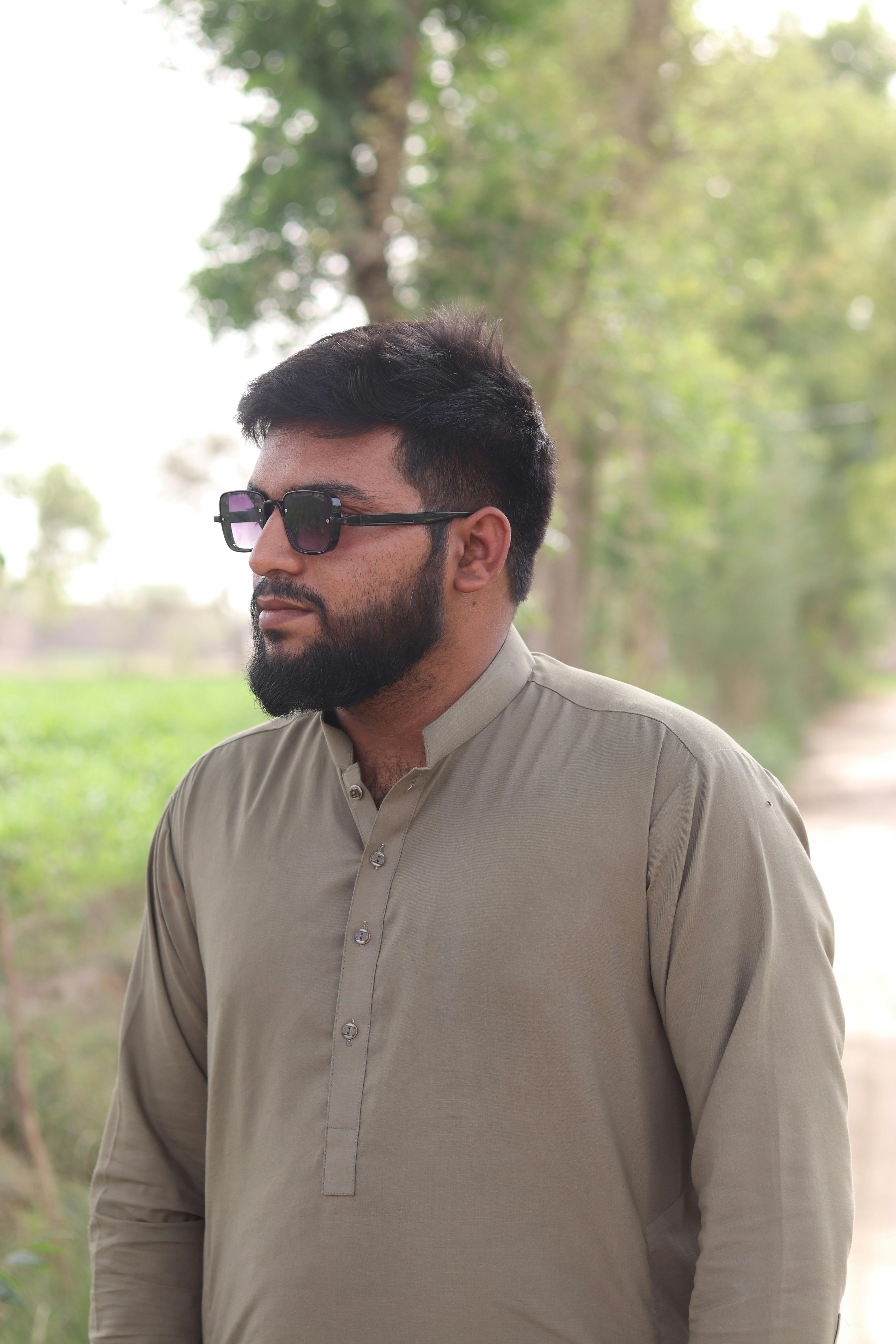 Adult man wearing sunglasses and traditional attire standing on a rural path surrounded by greenery.