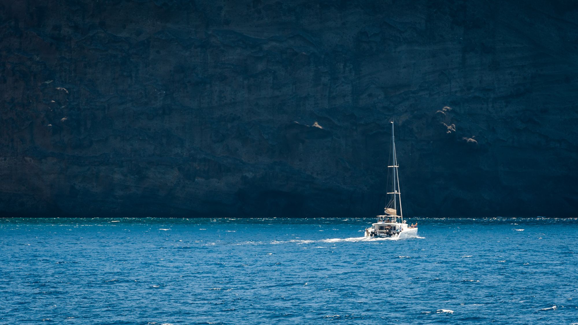 A sailing yacht under way on deep blue water beside a steep Mediterranean coastline