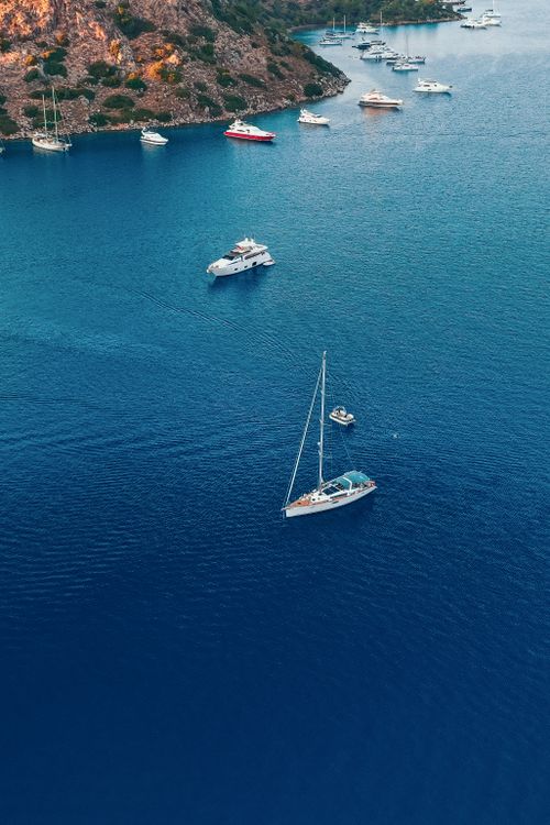 Free A tranquil aerial view of sailboats and yachts anchored in calm blue waters near a rocky coastline. Stock Photo