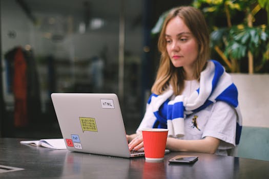 A young woman works on her laptop in a café setting, surrounded by greenery and a coffee cup.