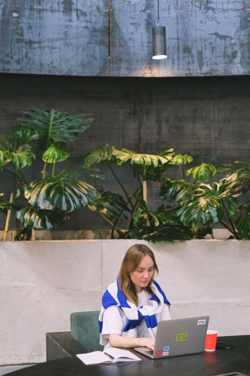 Free Woman using a laptop at a wooden table surrounded by plants, creating a calm workspace. Stock Photo