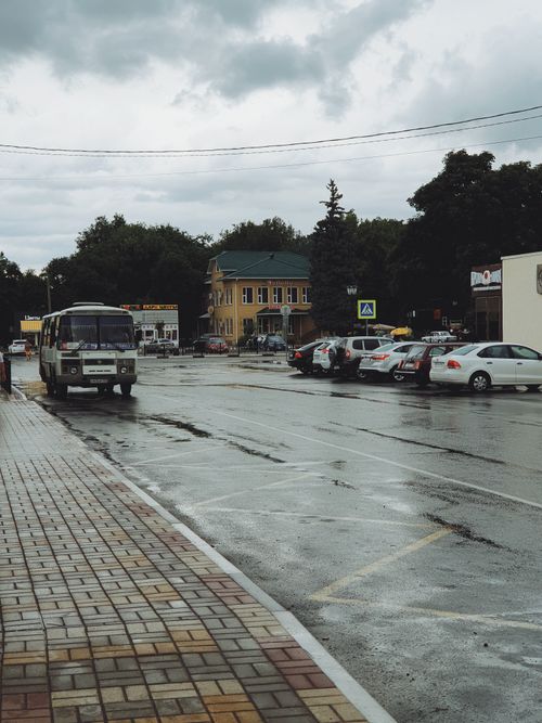 Free A rainy street scene showing parked cars, a bus, and buildings under a cloudy sky. Stock Photo