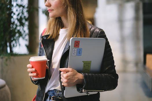 Young woman in a black leather jacket holding a laptop and takeaway coffee cup indoors.