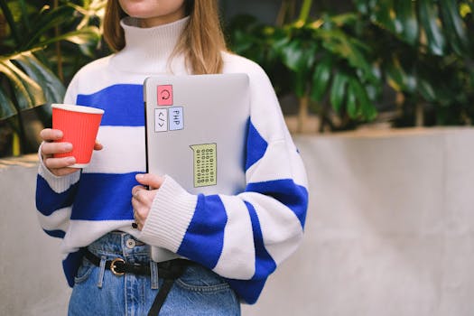 Woman in striped sweater holding laptop and coffee cup, exuding casual student life.