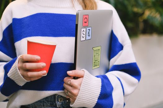 Person wearing striped sweater holding laptop with stickers and a red coffee cup indoors.