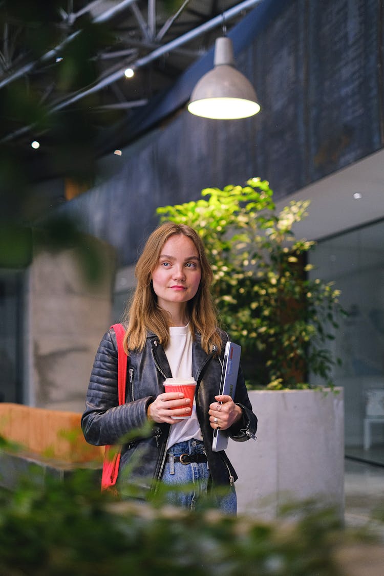 Woman Holding Laptop And Coffee Walking Down Hallway With Potted Plants